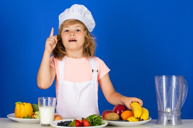 Portret letniego dziecka w czapce kucharskiej i fartuchu, robi sałatkę owocową i gotuje jedzenie w kuchni c