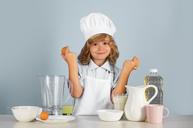 Portret letniego dziecka w czapce kucharskiej i fartuchu trzyma jajka, robi sałatkę owocową i gotuje jedzenie w