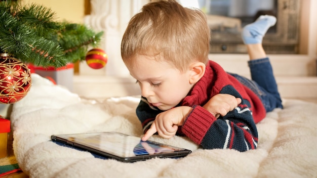 Portret małego chłopca leżącego na kocu obok choinki i grającego na cyfrowym tablecie