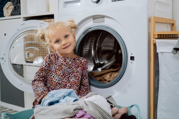 Portret małej dziewczynki o niebieskich oczach i blond włosach siedzącej przy otwartej pralce dziecko wyciąga ubrania z miski i wkłada je do bębna pracowita kreatywna pomocna córka