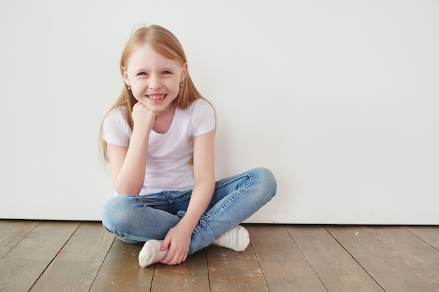Portret małej dziewczynki siedzącej na drewnianej podłodze w pobliżu białej ściany