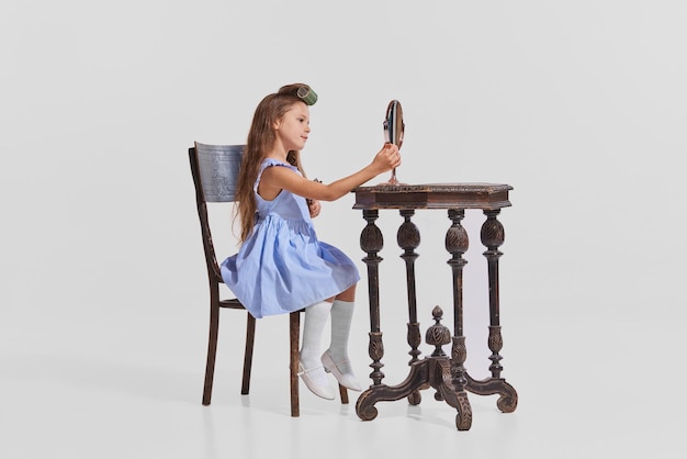 Portret małej dziewczynki siedzącej przy stole i robiącej makijaż patrząc na lustro na białym tle