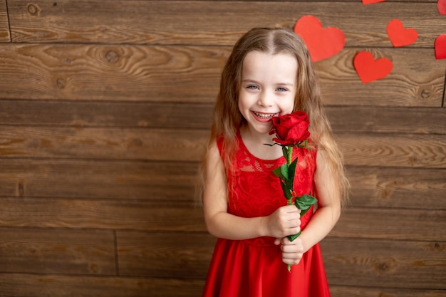 Portret małej dziewczynki w czerwonej sukience trzyma czerwoną różę na ciemnobrązowym drewnianym tle i uśmiecha się słodko koncepcja Walentynki puste miejsce na tekst