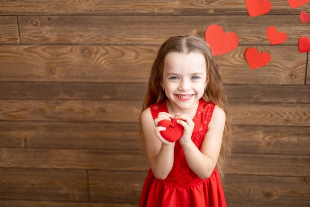 Portret małej dziewczynki w czerwonej sukience trzymającej czerwone zabawki na ciemnobrązowym drewnianym tle i słodko uśmiechającej się koncepcja Dnia Miłości pusta przestrzeń dla tekstu
