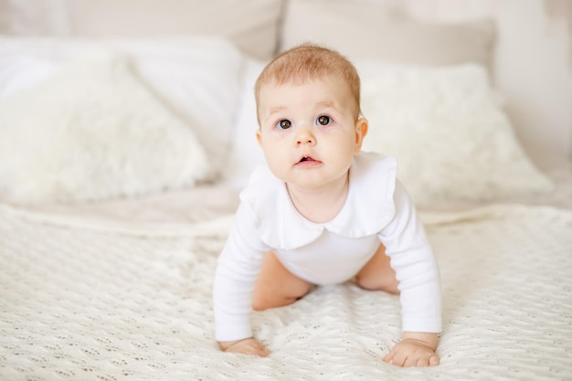 Portret małej dziewczynki w wieku sześciu miesięcy na łóżku w domu w jasnej sypialni uśmiecha się szczęśliwego noworodka w białym bawełnianym body czołgającym się