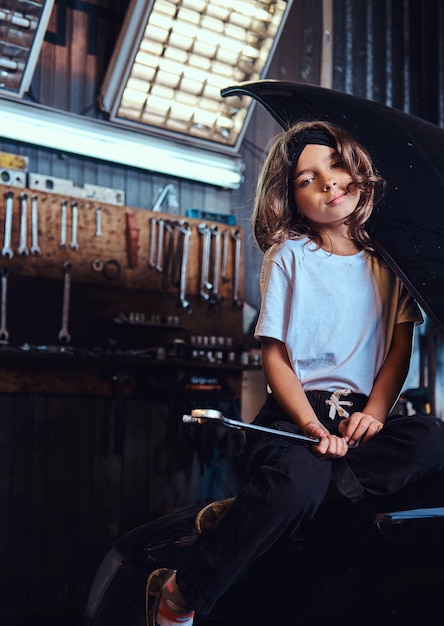 Portret małej pięknej dziewczyny z dużym narzędziem w ręce. Stoi w pobliżu samochodu w warsztacie samochodowym.