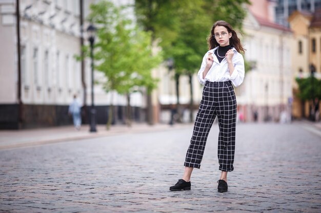 Portret małej pięknej stylowej dziewczynki z okularami przeciwsłonecznymi i krótkimi spodniami w kratę na miejskiej ulicy