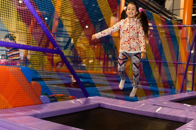 Portret małej słodkiej dziewczynki bawiącej się i skaczącej na trampolinie w pokoju zabaw dla dzieci, krytym placu zabaw. Aktywna dziewczyna bawiąca się w centrum sportowym.