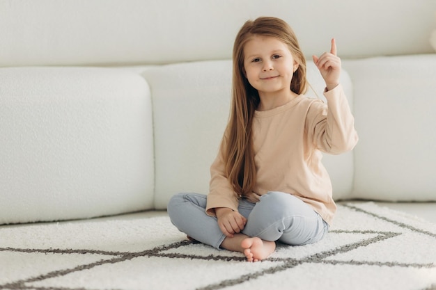 Portret małej słodkiej dziewczynki Siedzi na podłodze obok sofy Dziewczyna patrzy w kamerę i pokazuje palec w górę