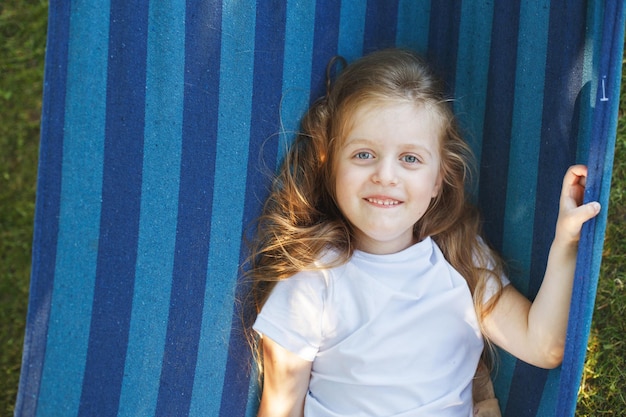 Portret małej słodkiej dziewczyny z długimi włosami, spoczywającej na hamaku w ogrodzie i uśmiechniętej