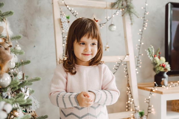 Portret Małej Wesołej Dziewczyny Z Ciemnymi Włosami Stojącej W Pobliżu Choinki.