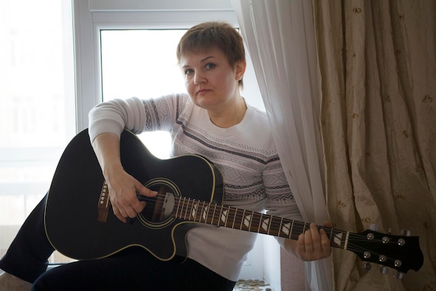 Portret marzycielskiej kobiety z gitarą w pobliżu okna w domu, zbliżenie