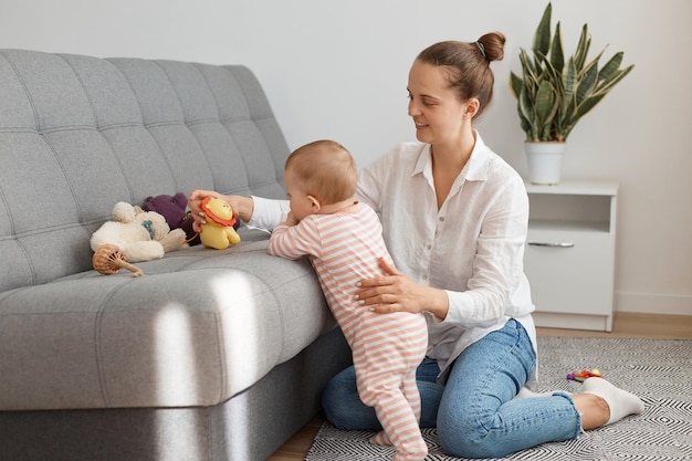 Portret matki i córki bawiące się razem siedząc na podłodze przy kanapie, mama uśmiechnięta radośnie, dziewczynka stojąca i wyciągająca rękę do kolorowych zabawek.