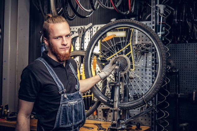 Portret mechanika rowerowego brodaty czerwony głowa w warsztacie z części rowerowych i koła na tle.