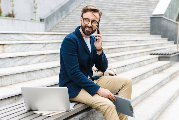 Portret Męski Biznesmen W Okularach, Trzymając Schowek I Rozmawia Przez Telefon, Siedząc Na ławce W Pobliżu Schodów