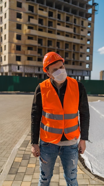 Portret męskiego pracownika budowlanego w masce medycznej i kombinezonie na tle domu w budowie