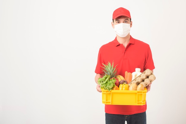 Portret mężczyzna w czerwień mundurze i twarzy masce trzyma świeżą żywność w plastikowym pudełku na białym tle, Bezpieczny domowy doręczeniowy pojęcie