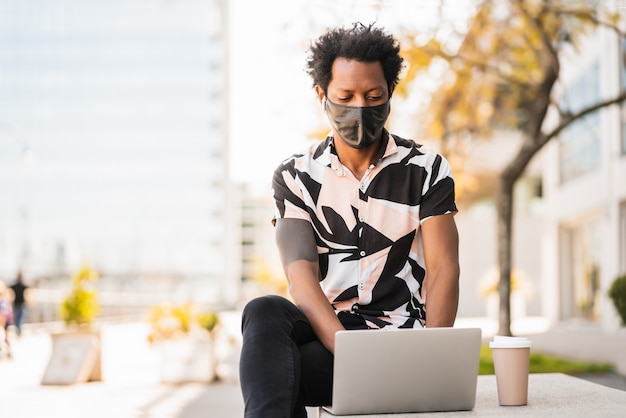 Portret mężczyzny afro turysta za pomocą swojego laptopa i nosząc maskę ochronną siedząc na zewnątrz. Koncepcja turystyki. Nowa koncepcja normalnego stylu życia.