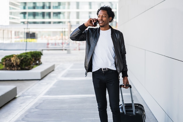 Portret mężczyzny afro turystycznego rozmawiającego przez telefon i niosącego walizkę podczas spaceru na świeżym powietrzu po ulicy