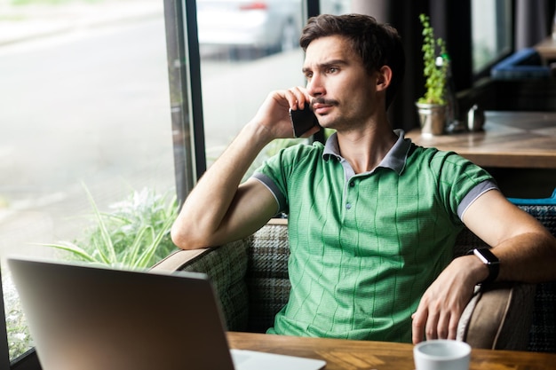 Portret mężczyzny freelancer ubrany w zieloną koszulkę, mówiący telefon komórkowy, omawiający problemy, odwracając wzrok, rozmawiając z partnerem biznesowym. Strzał kryty w pobliżu tła kawiarni z dużym oknem