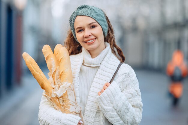 Portret miasta na rozmytym tle. Młoda atrakcyjna kobieta w białym garniturze z dzianiny iz ciemną torbą stoi i uśmiecha się słodko, ściskając obiema rękami kilka bagietek z chlebem.