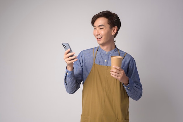 Portret młodego azjatyckiego mężczyzny noszącego fartuch, trzymającego smartfona i papierowy kubek na białym tle