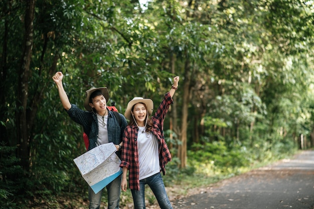 Portret młodego azjatyckiego przystojny mężczyzna z plecakiem i kapeluszem trekkingowym i ładną dziewczyną stojącą i sprawdzającą kierunek na papierowej mapie podczas chodzenia po leśnym szlaku, koncepcja podróży plecakiem