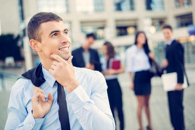 Portret młodego biznesmena myślącego stojącego przed budynkiem biurowym oddzielonym od reszty zespołu biznesowego.