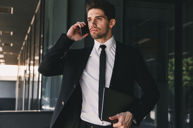 Portret młodego biznesmena ubranego w formalny garnitur spaceru na zewnątrz budynku ze szkła i rozmawia przez telefon komórkowy