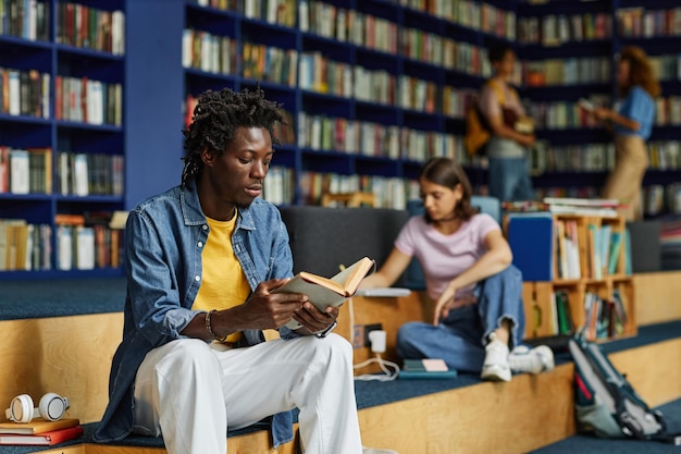 Portret młodego czarnego mężczyzny czytającego książkę w salonie bibliotecznym ze studentami w przestrzeni kopii w tle