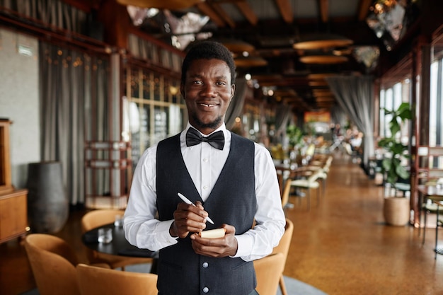 Portret młodego czarnego mężczyzny jako kelner w luksusowej restauracji uśmiechający się do kamery i trzymający notatnik gotowy do przyjmowania zamówień.