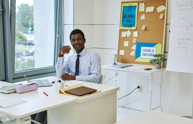 Portret młodego czarnego nauczyciela uśmiechającego się do kamery siedząc przy biurku w pustej klasie kopiuj przestrzeń