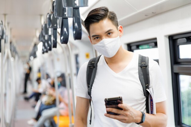 Portret młodego dorosłego mężczyzny z Azji w masce medycznej, stojącego, trzymającego smartfon i patrzącego na kamerę w pociągu Skytrain z rozmytym tłem Skytrain. Nowa koncepcja normalnego stylu życia