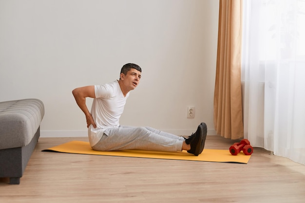 Portret młodego dorosłego przystojnego sportowca siedzącego na macie, wykonującego ćwiczenia rozciągające jogi na siłowni, cierpiących na ból pleców dotykający pleców z bólem mięśni dłoni