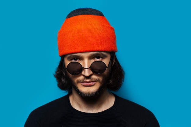 Portret Młodego Faceta Pewnie Sobie Okulary, Pomarańczowy Kapelusz Beanie I Czarny Sweter Na Tle Koloru Niebieskiego.