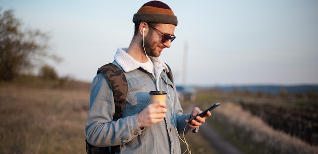 Portret młodego faceta, trzymając kubek gorącej kawy i smartphone z zewnątrz słuchawki