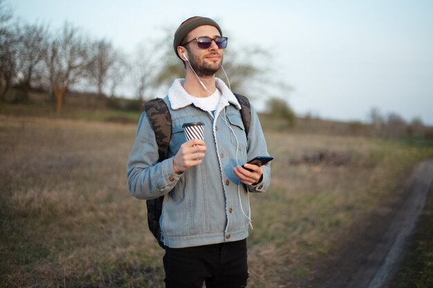 Portret młodego faceta, trzymając kubek gorącej kawy i smartphone z zewnątrz słuchawki