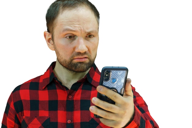 Portret młodego faceta w czerwonej koszuli w kratę na białym tle iz telefonem w dłoniach Brodaty mężczyzna patrzy na telefon ze zdziwionym wyrazem twarzy