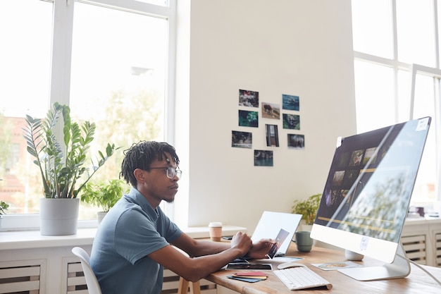 Portret młodego fotografa afroamerykańskiego przy użyciu komputera przy biurku w biurze domowym z oprogramowaniem do edycji zdjęć na ekranie, kopia przestrzeń