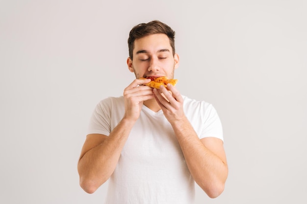 Portret młodego głodnego mężczyzny z gryzącym pyszny kawałek pizzy, z zamkniętymi oczami z przyjemności na na białym tle. Strzał studio przystojny student płci męskiej jedzenie smaczne jedzenie.