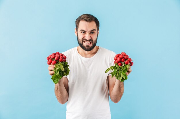 Portret Młodego, Gniewnego, Zniesmaczonego Brodatego Mężczyzny W Koszulce, Stojącego Na Białym Tle Nad Niebieską ścianą, Pokazującego świeży Czerwony Kolor