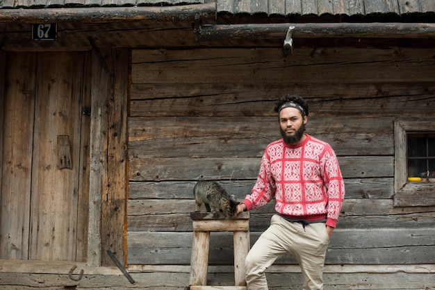 portret młodego hipstera z brodą i kotem przed drewnianym domem