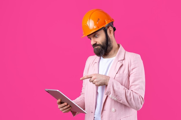 Portret Młodego Inżyniera Budowlanego W Pomarańczowym Kasku W Różowej Kurtce Stojącego Na Czerwonym Tle Studia Mężczyzna Z Tabletem W Dłoniach