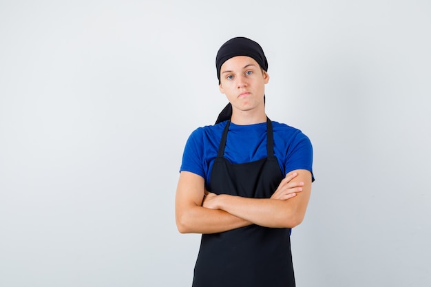 Portret młodego kucharza z założonymi rękoma w koszulce, fartuchu i patrzącym na zamyślony widok z przodu