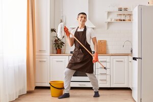 Zdjęcie portret młodego mężczyzny myjącego podłogę w kuchni, słuchającej muzyki i tańczącej, trzymając w rękach mopa, sprzątając swoje mieszkanie, patrząc na kamerę w ubraniu casual i brązowym fartuchu