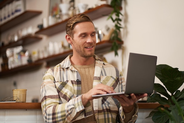 Portret młodego mężczyzny pracującego online jako freelancer siedzący w kawiarni wykonujący swój projekt na laptopie pijący
