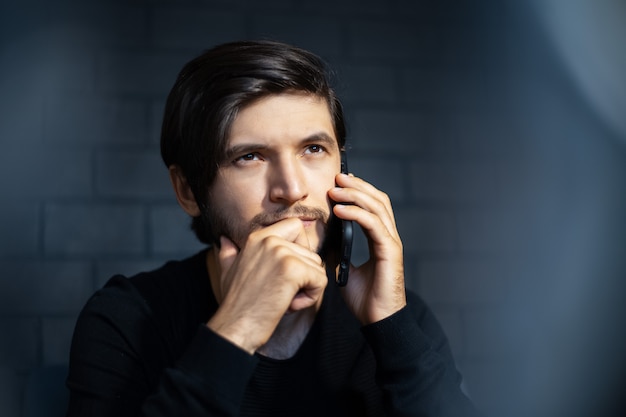 Portret młodego mężczyzny, rozmawia przez smartfona. Na tle czarnej cegły ściany.