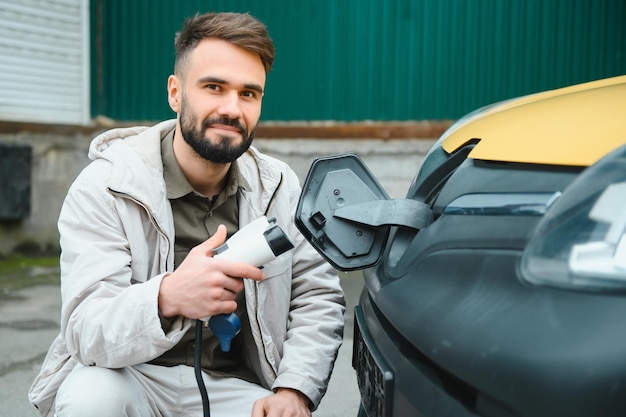 Portret młodego mężczyzny stojącego z kablem ładującym w pobliżu stacji ładującej