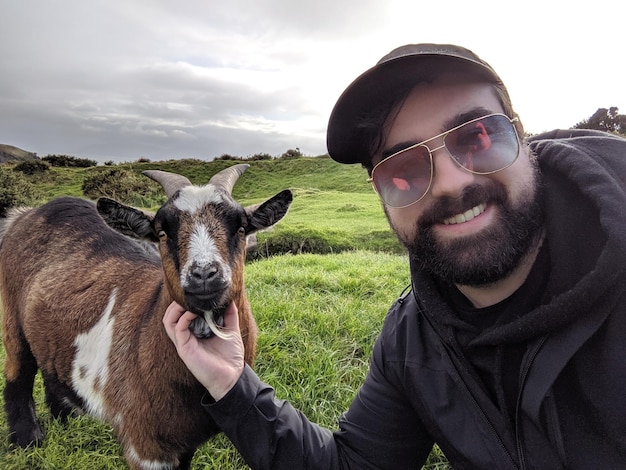 Zdjęcie portret młodego mężczyzny w okularach przeciwsłonecznych i pozującego z kozą w irlandii