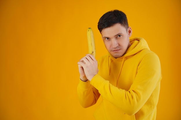 Portret młodego mężczyzny z bananem na żółtym tle Zbliżenie przystojnego faceta w żółtej bluzie z kapturem bawi się owocami, wyobrażając sobie go jako broń